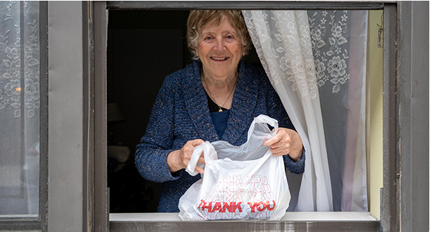 an older woman standing in a window with a lace curtain holds a plastic bag containing a meal and smiles