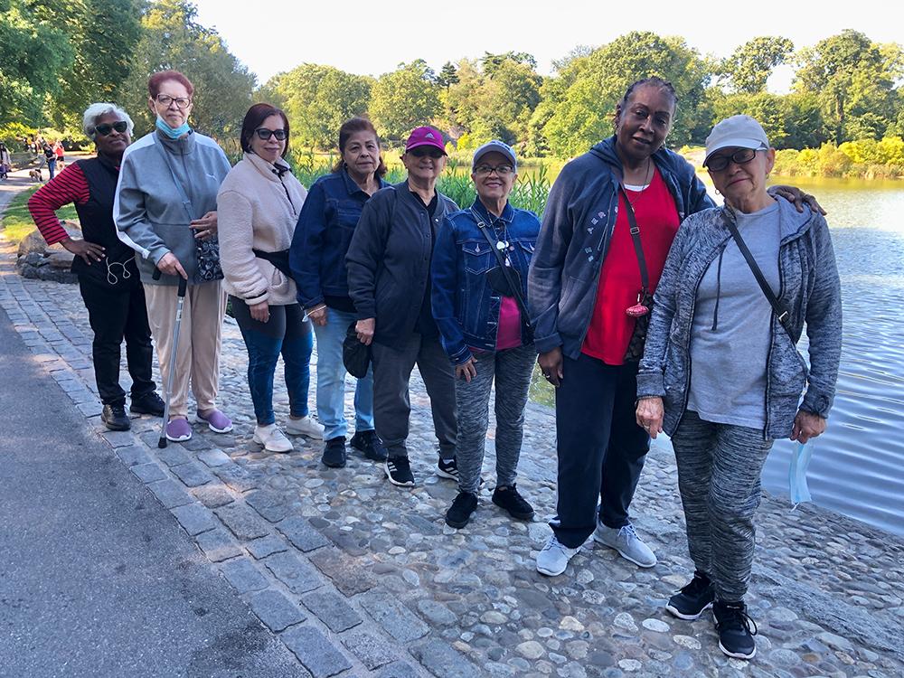 Several women stand in a row in front of the Harlem Meer on a sunny day