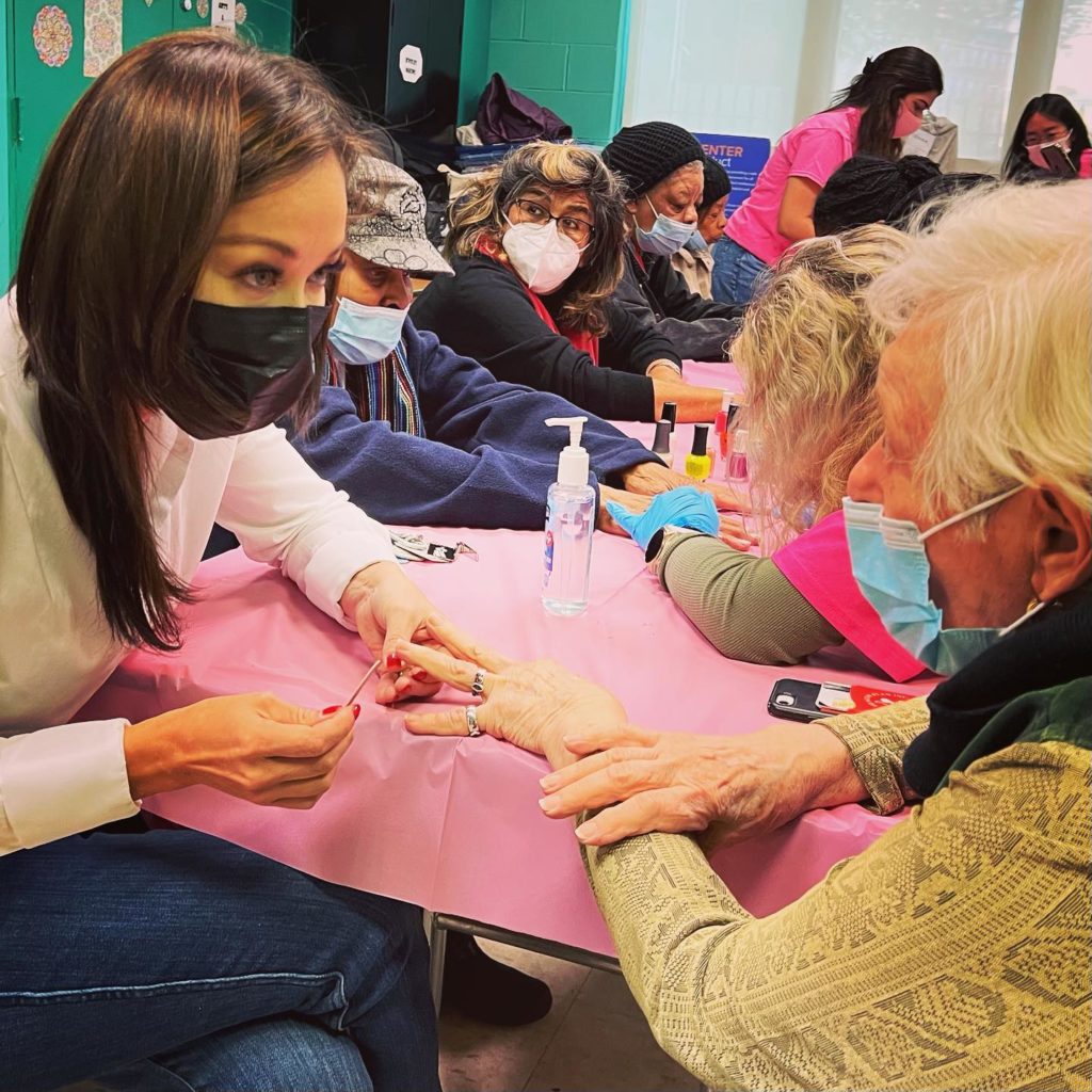 A young woman wearing a black face mask leans forward and makes eye contact with an older woman while painting her nails
