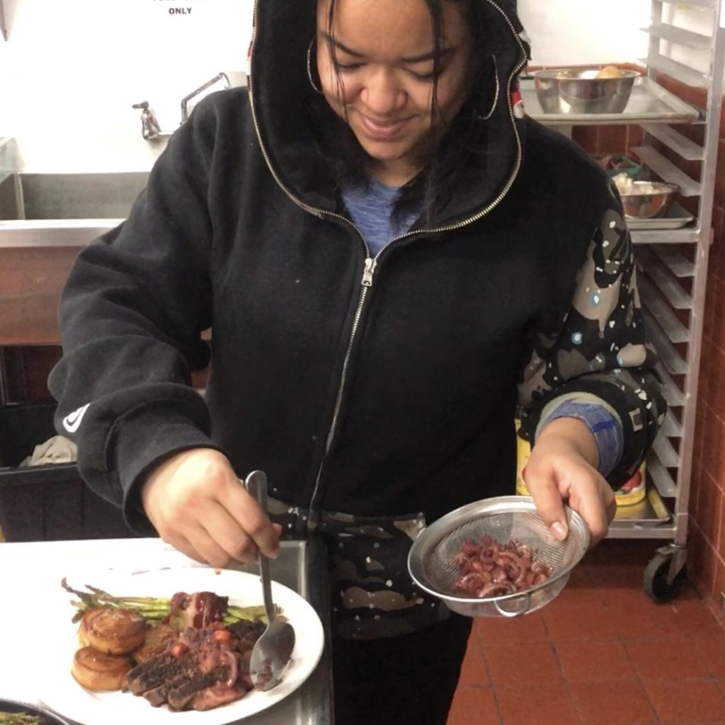 A young woman uses a spoon to arrange diced vegetables on top of steak in an industrial kitchen