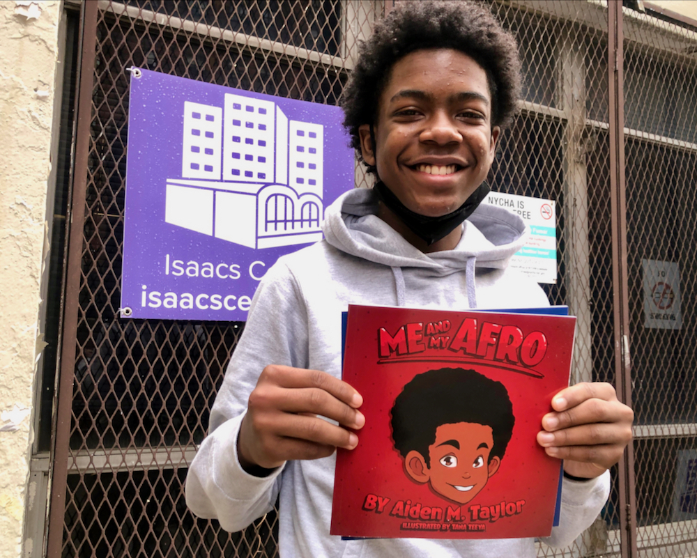 A young man stands in front of an Isaacs Center sign holding a copy of ME AND MY AFRO