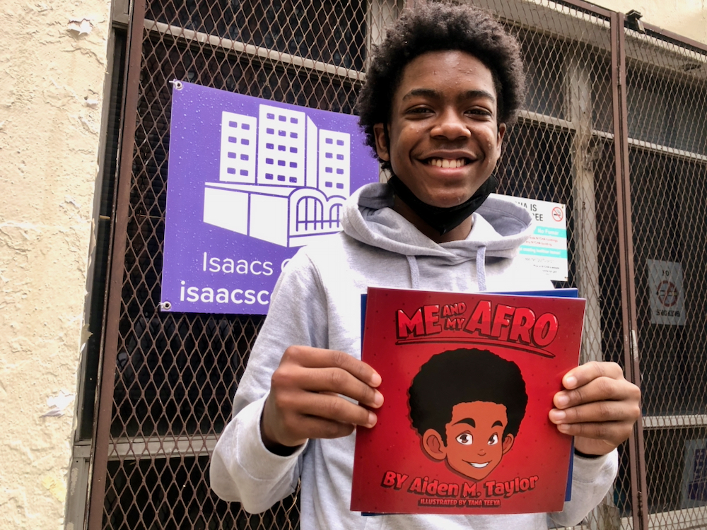 A young man stands in front of an Isaacs Center sign holding a copy of ME AND MY AFRO