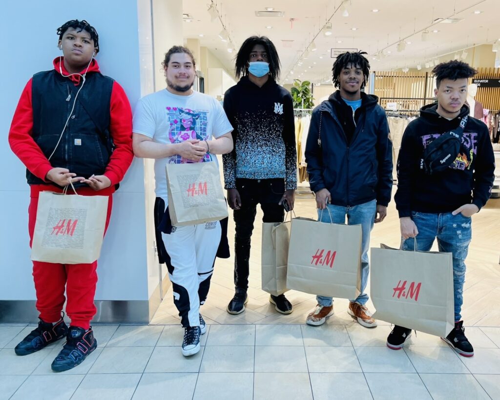 Five young men stand in front of a store in a mall holding shopping bags