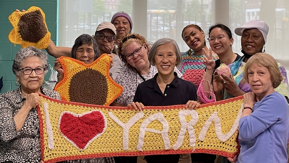 Several people stand behind a crocheted banner reading I Heart Yarn while some hold up large crochet flowers