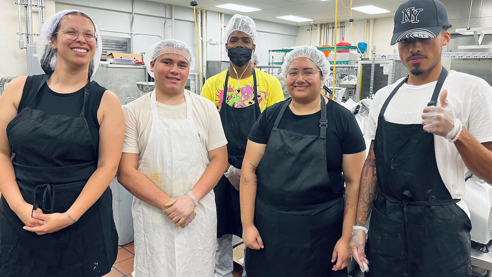 Five people in aprons and hairnets pose in a commercial kitchen
