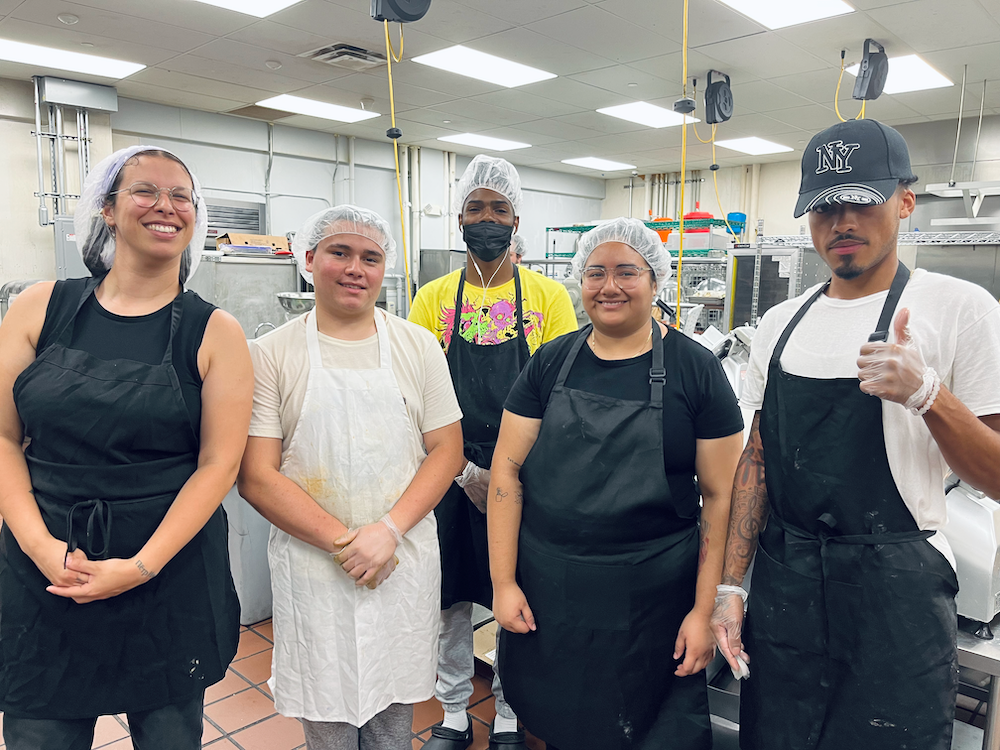 Five people in aprons and hairnets pose in a commercial kitchen
