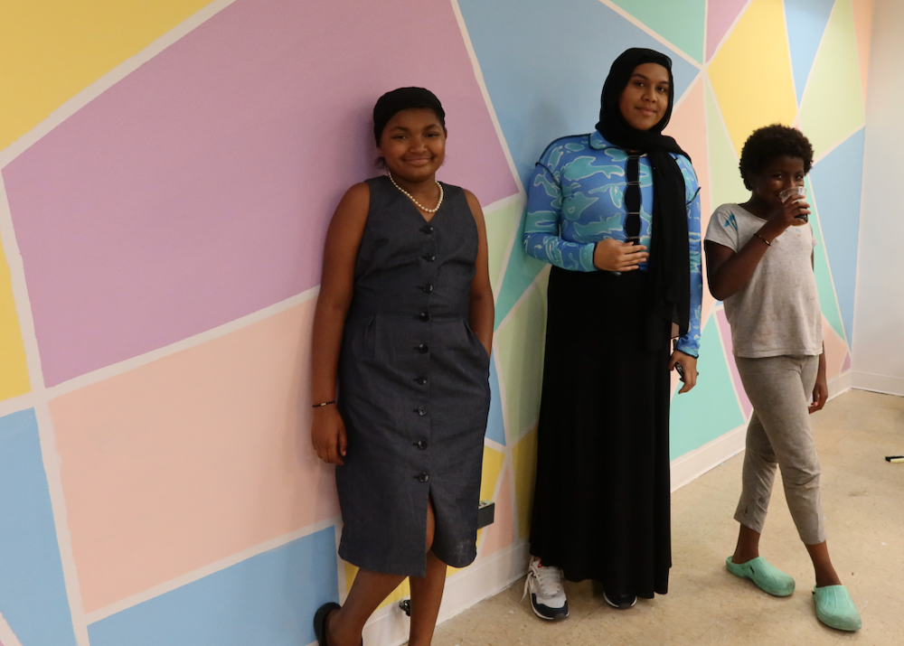Three young people stand in front of a mural of irregular blocks of peach pink blue and yellow