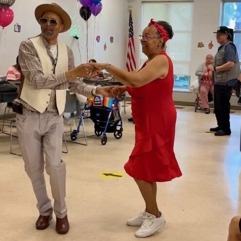 Two older adults hold hands and dance while others watch in a room decorated with balloons and colored lights