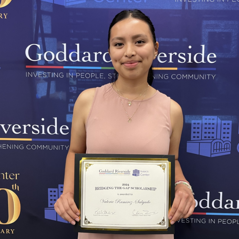 A young woman poses in front of a Goddard/Isaacs Center backdrop while holding her scholarship certificate