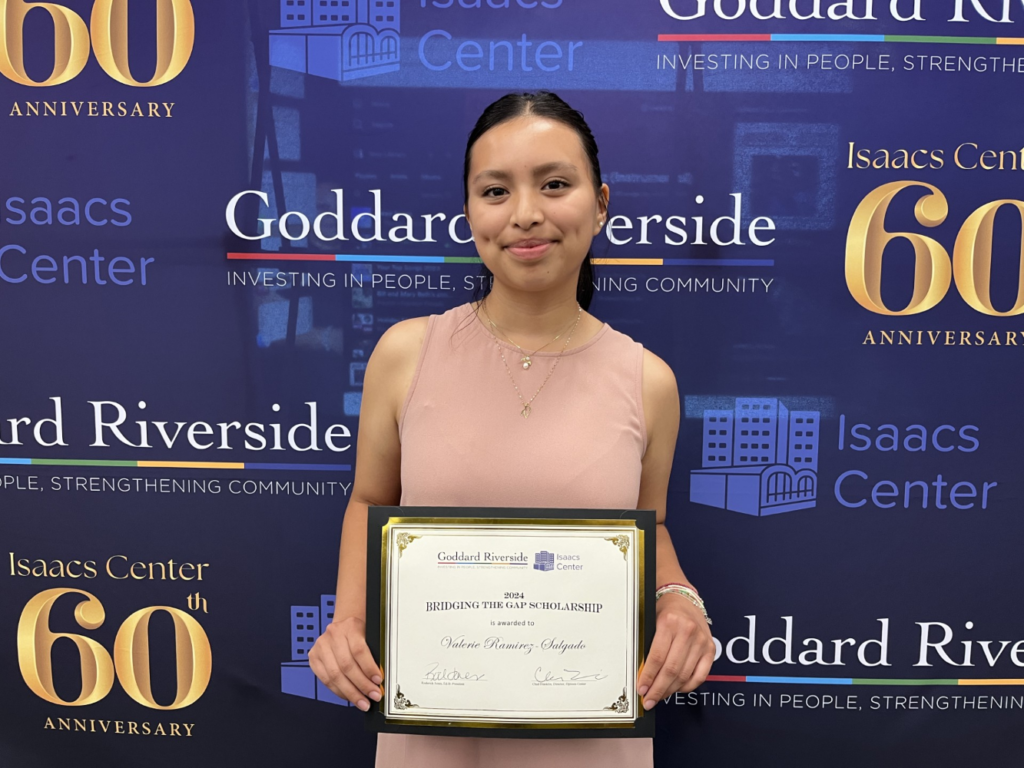 A young woman holds a scholarship certificate while standing in front of a Goddard/Isaacs Center backdrop