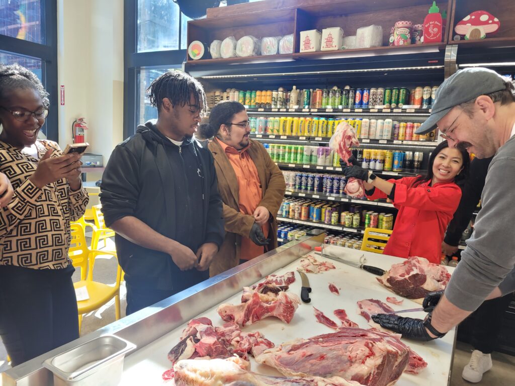 Four people standing around a butcher working on a piece of beef while a person holds a bone up