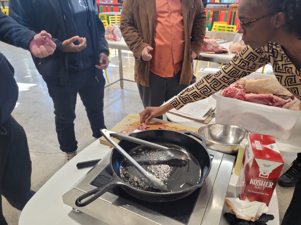 People standing over a used skillet grabbing pieces of steak