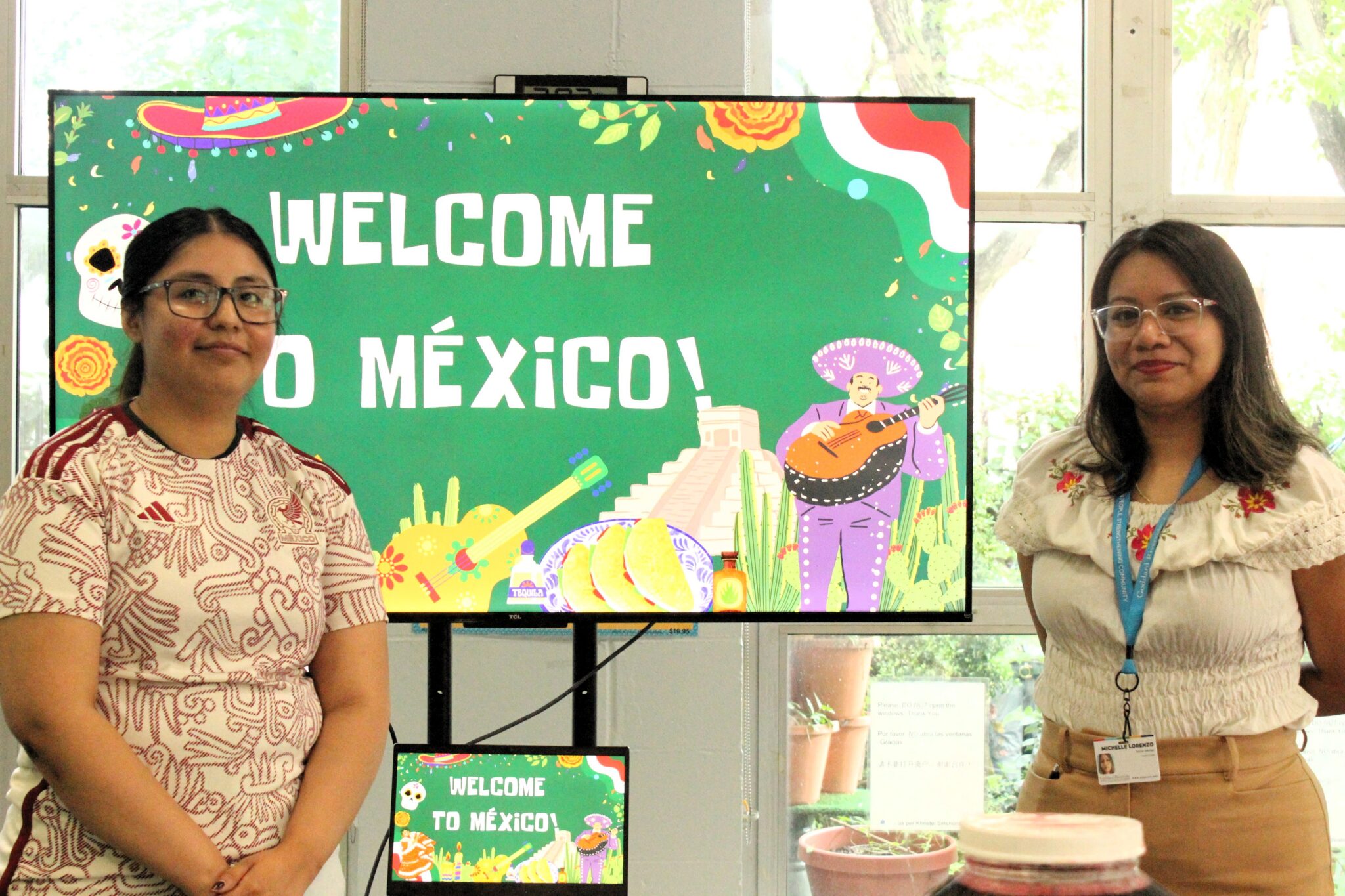 Two women stand flanking a video screen with the words Welcome to Mexico and a colorful gold and green design
